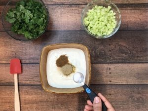 adding teaspoon sugar to yogurt in bowl with chopped herbs and cucumber on the side