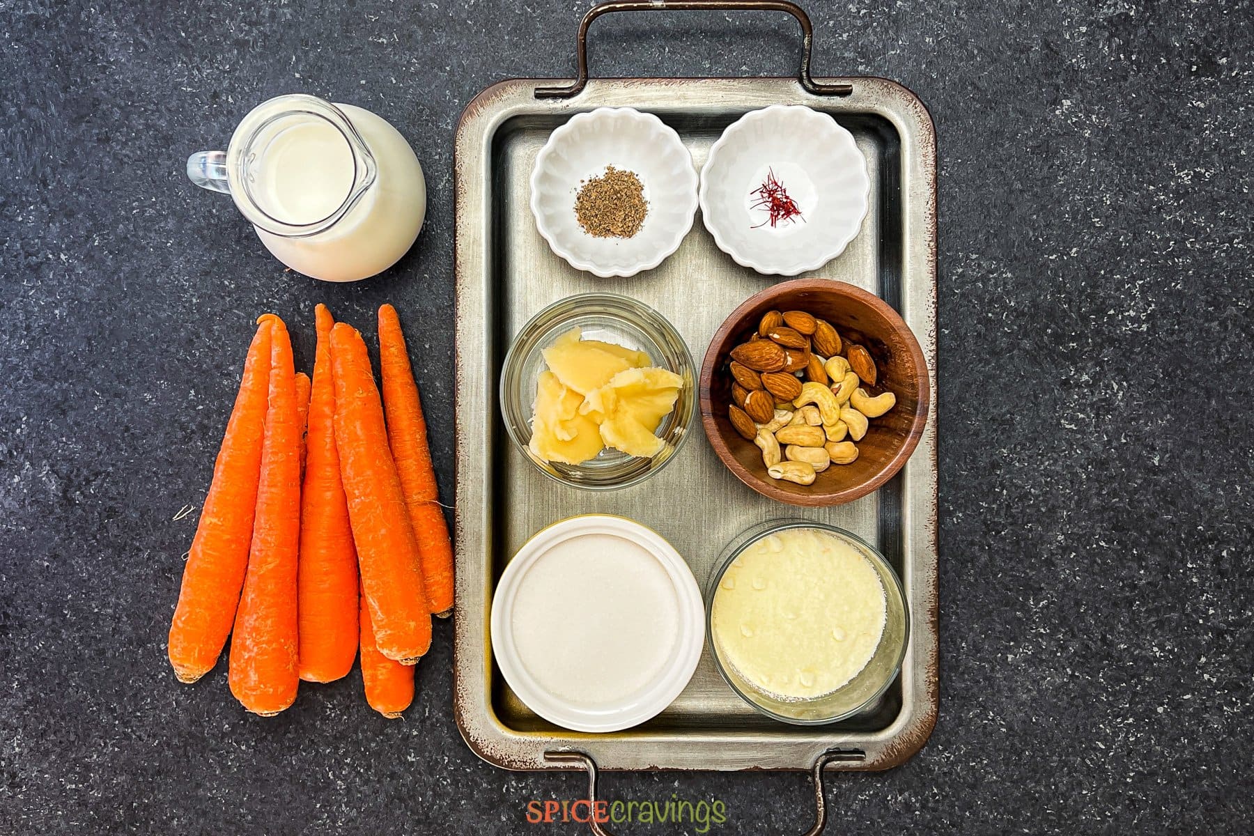 Carrots, milk, nuts, cardamom powder, milk powder and sugar on metal tray