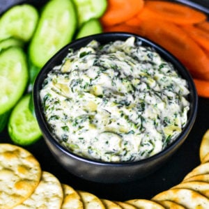 Spinach artichoke dip served in a black bowl with veggies and crackers around