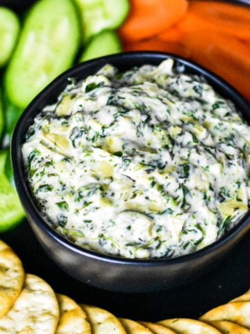 Spinach artichoke dip served in a black bowl with veggies and crackers around