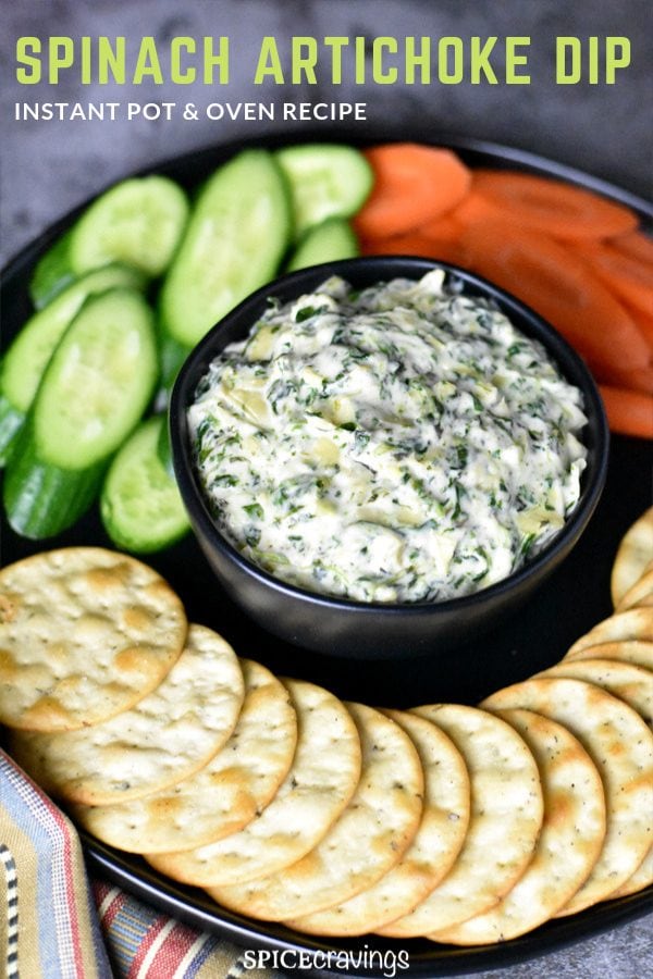 A bowl of spinach artichoke dip served with crackers, carrots and cucumbers