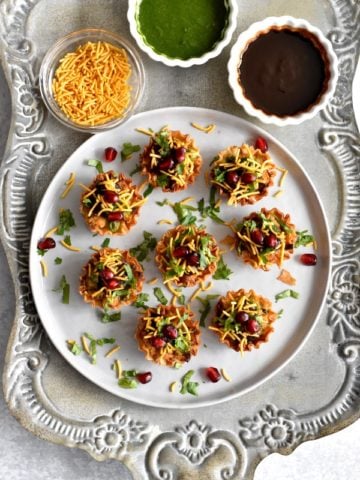 samosa chaat phyllo cups garnished with cilantro, pomegranate seeds and thin sev on white plate with chutneys in small bowls