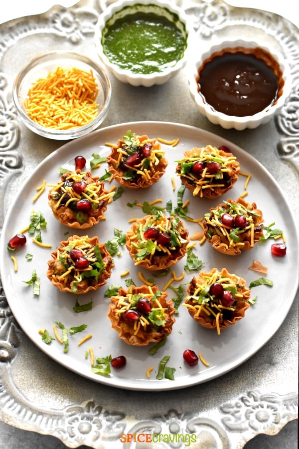 samosa chaat phyllo cups garnished with cilantro, pomegranate seeds and thin sev on white plate with chutneys in small bowls