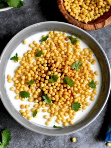 spiced yogurt condiment with boondi and cilantro in gray bowl with boondi in bowl on side