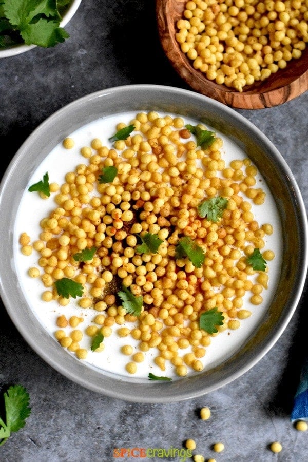 spiced yogurt condiment with boondi and cilantro in gray bowl with boondi in bowl on side