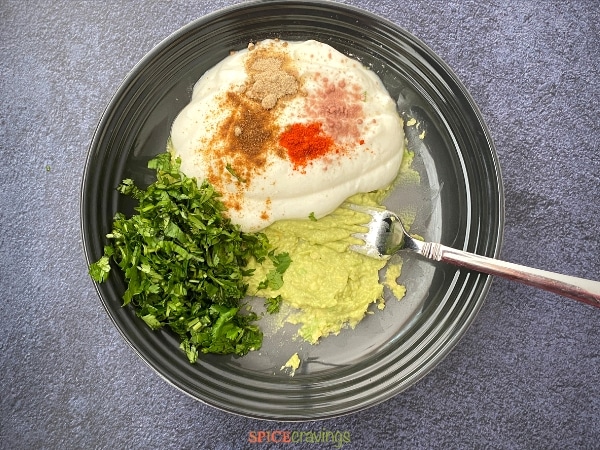 mashed avocado, yogurt, indian spices, chopped cilantro in stoneware bowl with fork