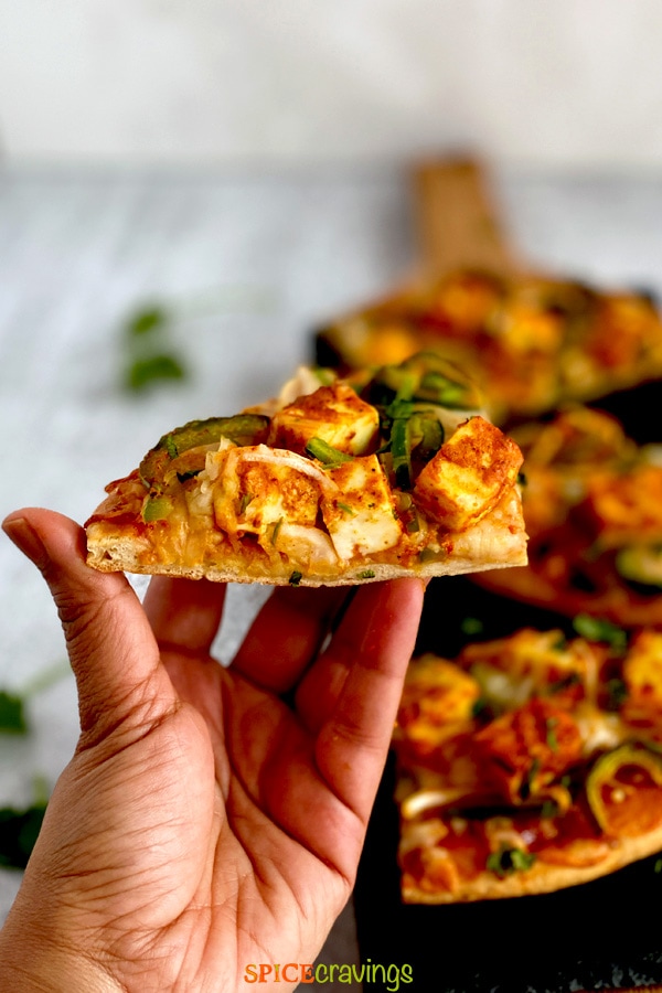 hand holding indian flatbread pizza with marinated paneer cheese and veggies on black board