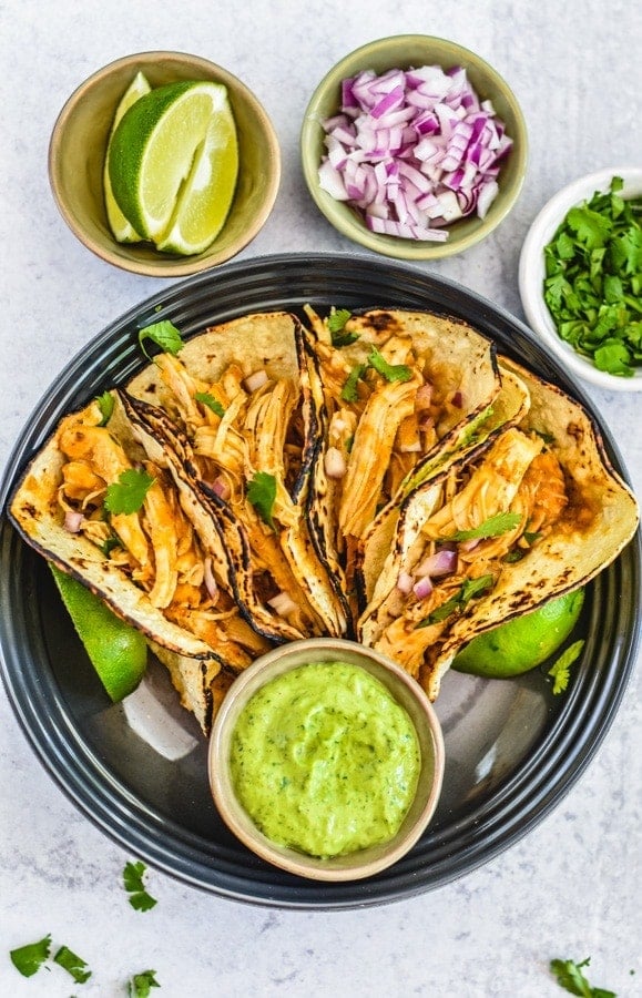 four corn tortillas filled with chicken tinga with avocao dressing on black plate with sliced lime, chopped red onion and cilantro