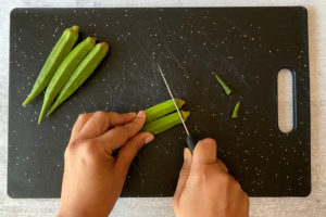 two hands cutting stems off fresh okra on black cutting board