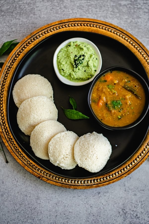 steamed idli on black plate with coconut chutney and sambar in two bowls