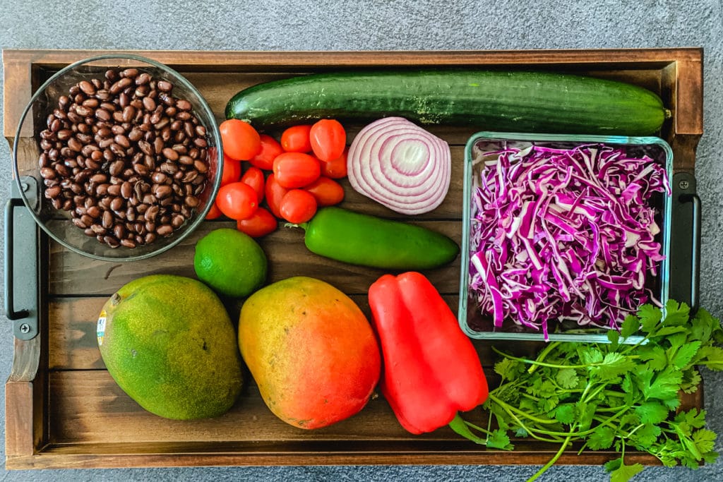 black beans, tomatoes, cucumber, red onion, sliced red cabbage, lime, mangoes, pepper, cilantro on wooden board