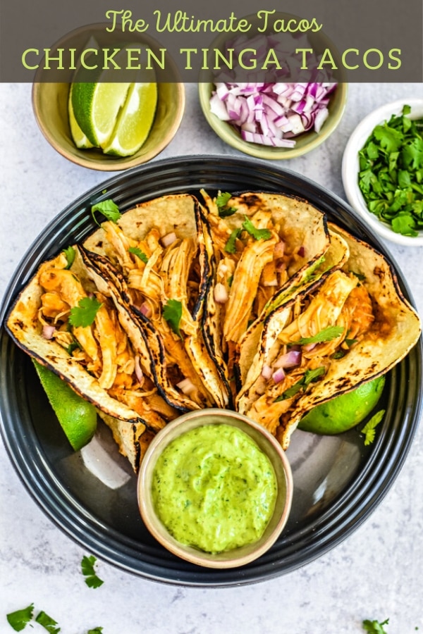 four corn tortillas filled with chicken tinga with avocao dressing on black plate with sliced lime, chopped red onion and cilantro