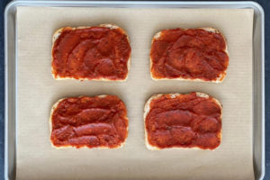 tomato sauce smeared on four slices of bread on parchment-lined baking sheet