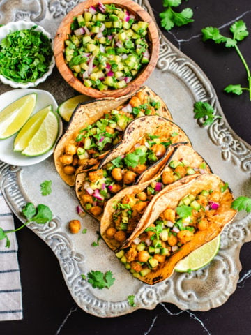 chana masala tacos with cucumber salsa on silver serving platter with lime wedges and cucumber salsa on side