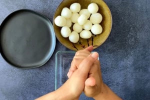two hands squeezing sugar syrup from rasgulla balls with extra rasgulla in brown bowl