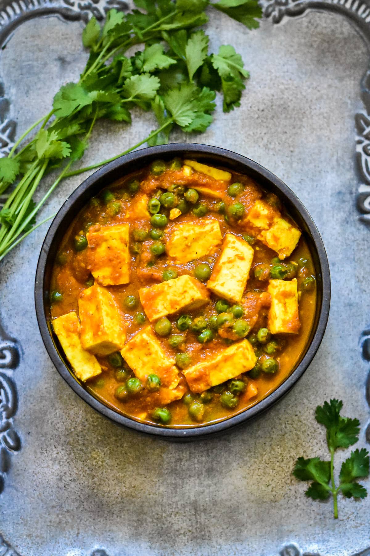 paneer and pea curry in black bowl with cilantro sprigs on silver platter