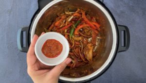 hand holding small bowl with Indian pickle juice over peppers, onions and aromatics in instant pot