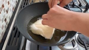 two hands placing piece of instant batura dough in hot oil on the stove
