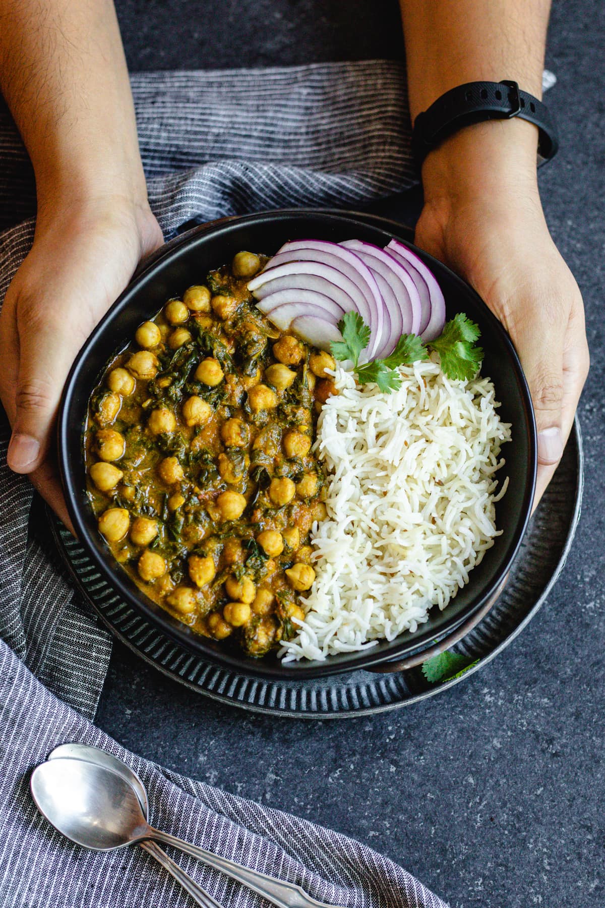 two hands holding chana saag and cumin rice with sliced red onions in black bowl