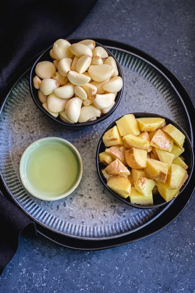 Two bowls of peeled garlic and ginger with a small bowl of oil on metal plate