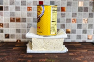 A block of tofu with paper towels underneath and on top, with a can pressing down on top