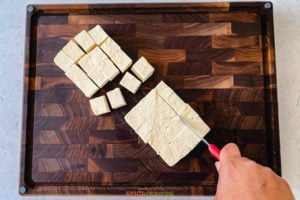 A hand slicing tofu into cubes on a cutting board