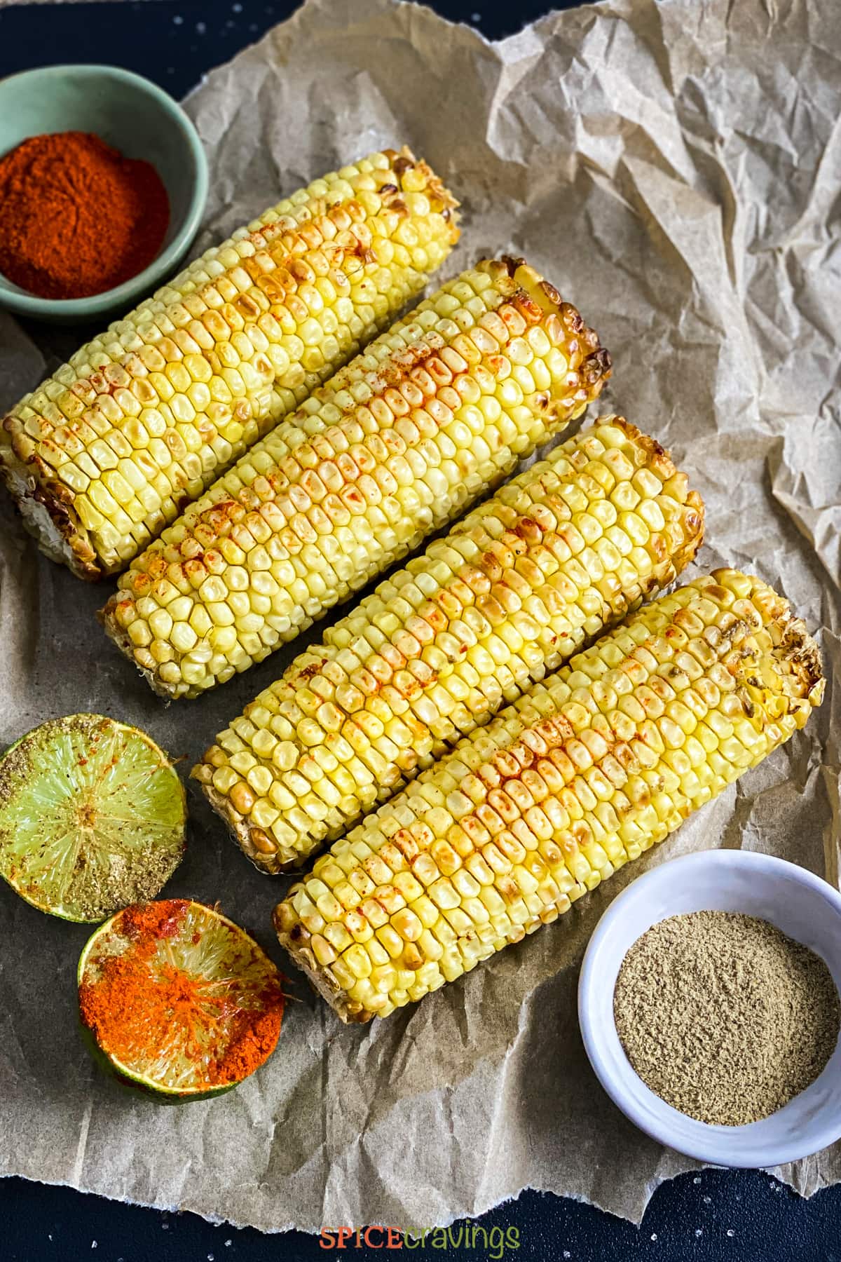 4 ears of roasted corn next to lime halves covered in spices