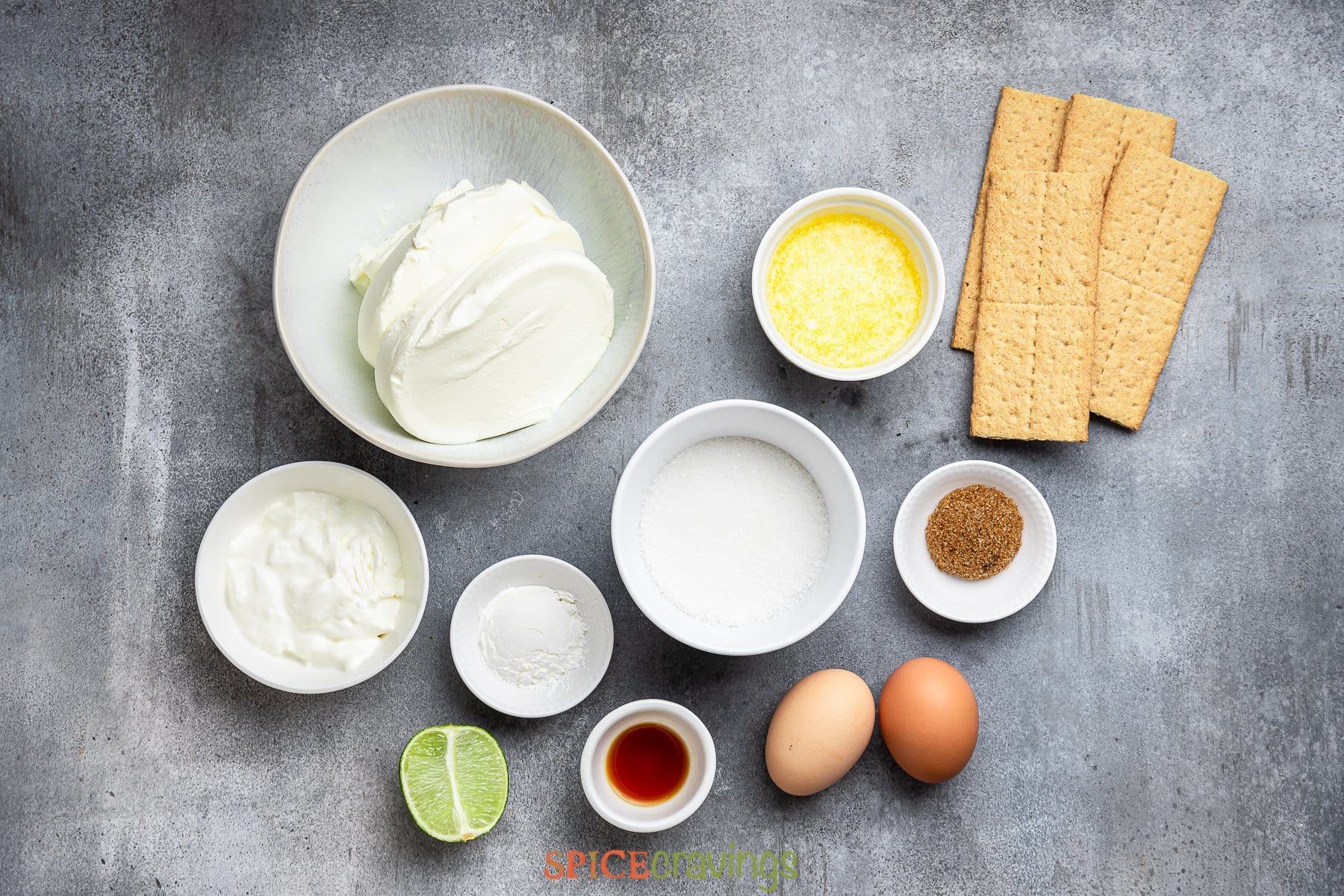 overhead of cheesecake ingredients on gray background including crackers, cream cheese and eggs