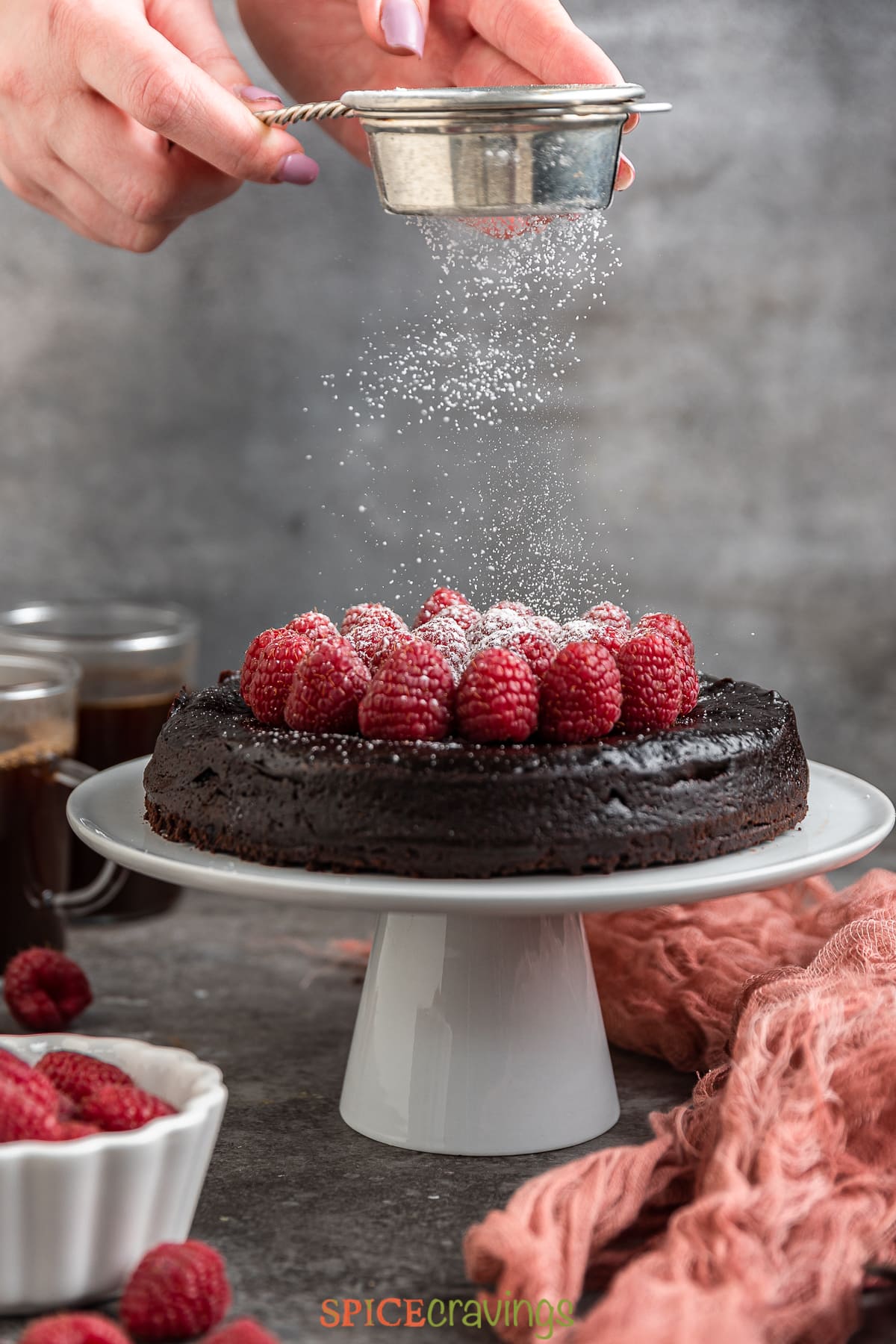 dusting powdered sugar over flourless chocolate cake topped with raspberries on cake stand