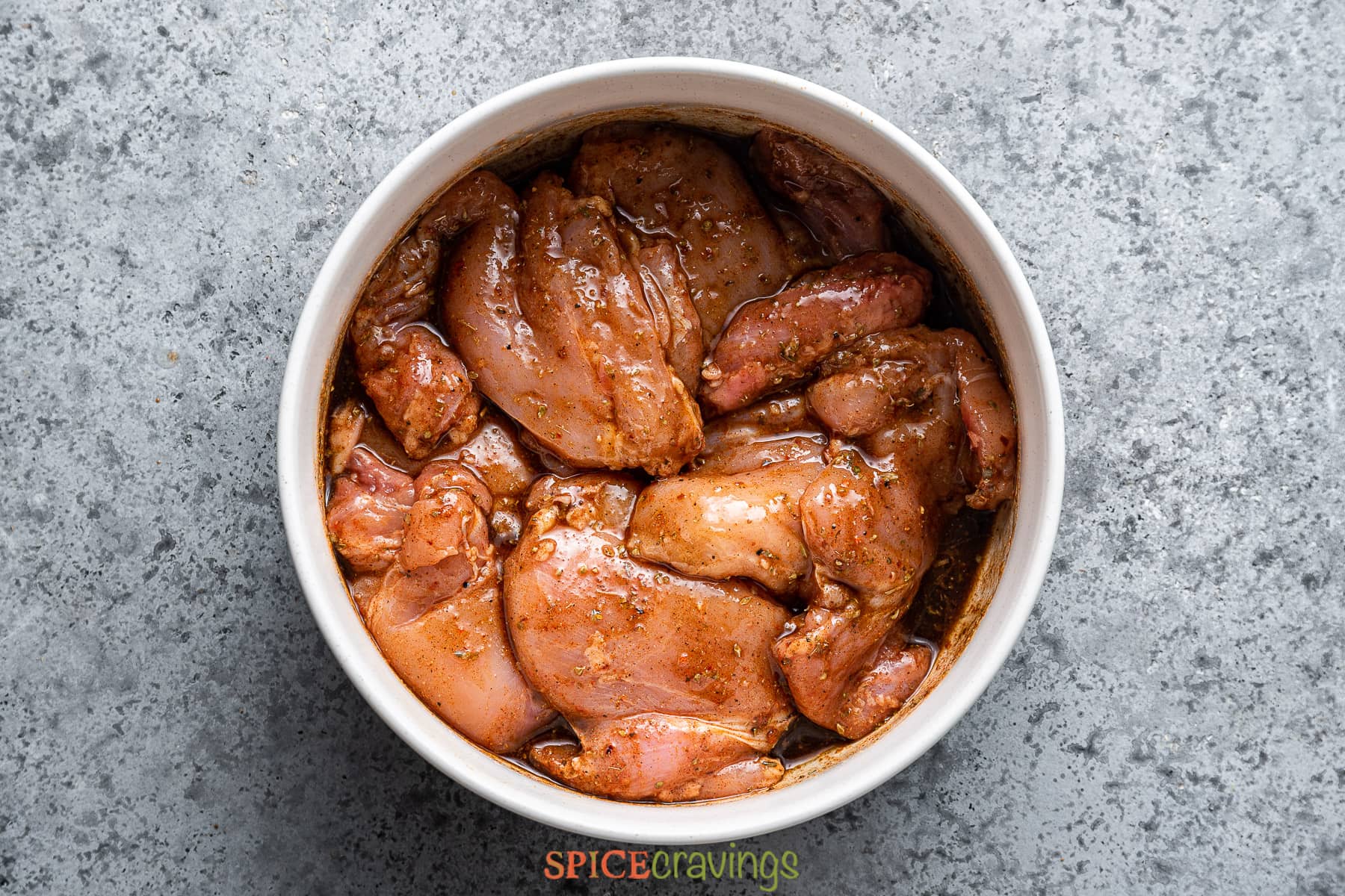 chicken thighs marinating in Greek herbs and spices in white bowl