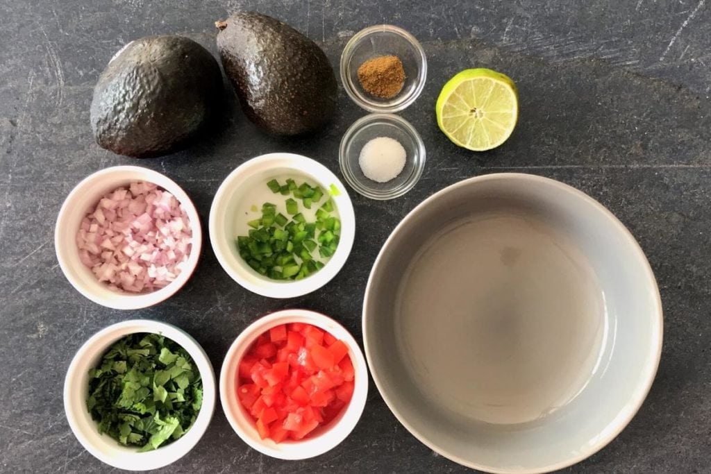 Avocado, chopped onion, tomato, cilantro among other Guacamole Ingredients on grey board