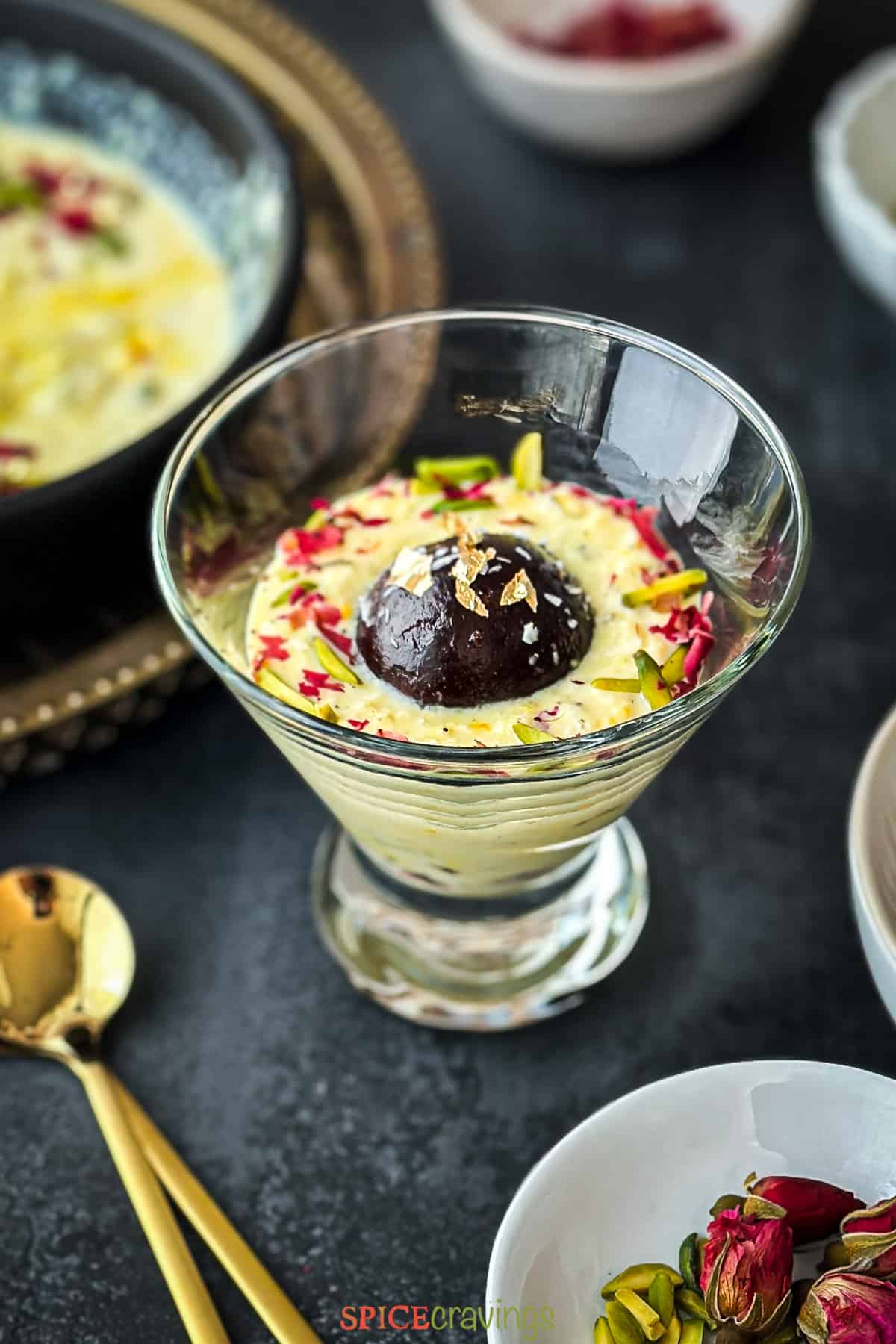 Black Gulab jamun dipped in a glass of rabdi and garnished with rose and pistachios