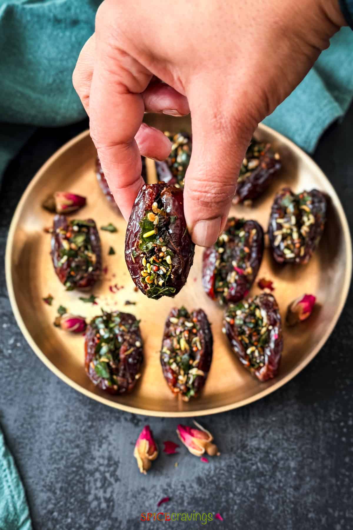Hand holding a single stuffed date paan against a background of a brass plate with dates paan and dried rose buds