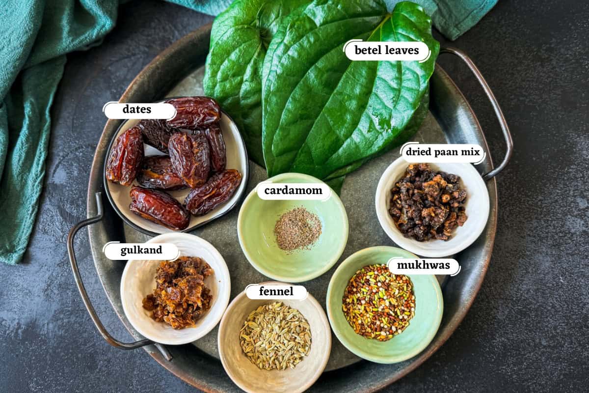 A colorful display of Indian ingredients on metal tray including dates, dried paan, gulkand, cardamom, fennel, and betel leaves for making dates paan.
