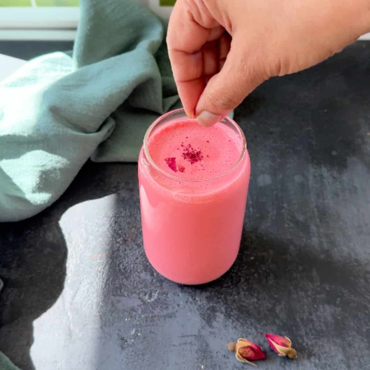 Sprinkling crushed dried rose petals on top of a glass with pink rose lassi