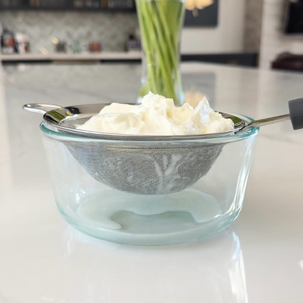 Straining Greek yogurt in a fine-mesh sieve over a glass bowl to create thick hung curd for Rose Shrikhand.