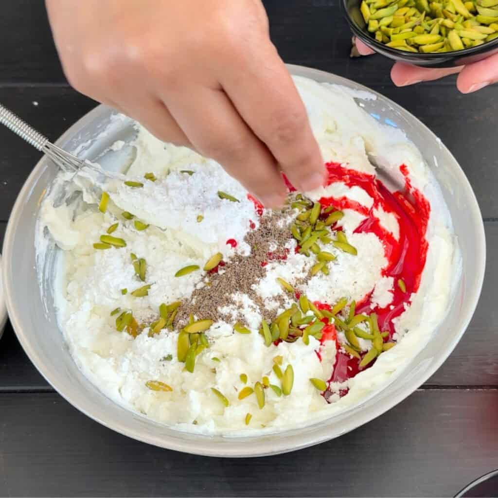 Adding rose syrup, cardamom powder, sugar, and slivered pistachios to the thickened yogurt base in a mixing bowl.