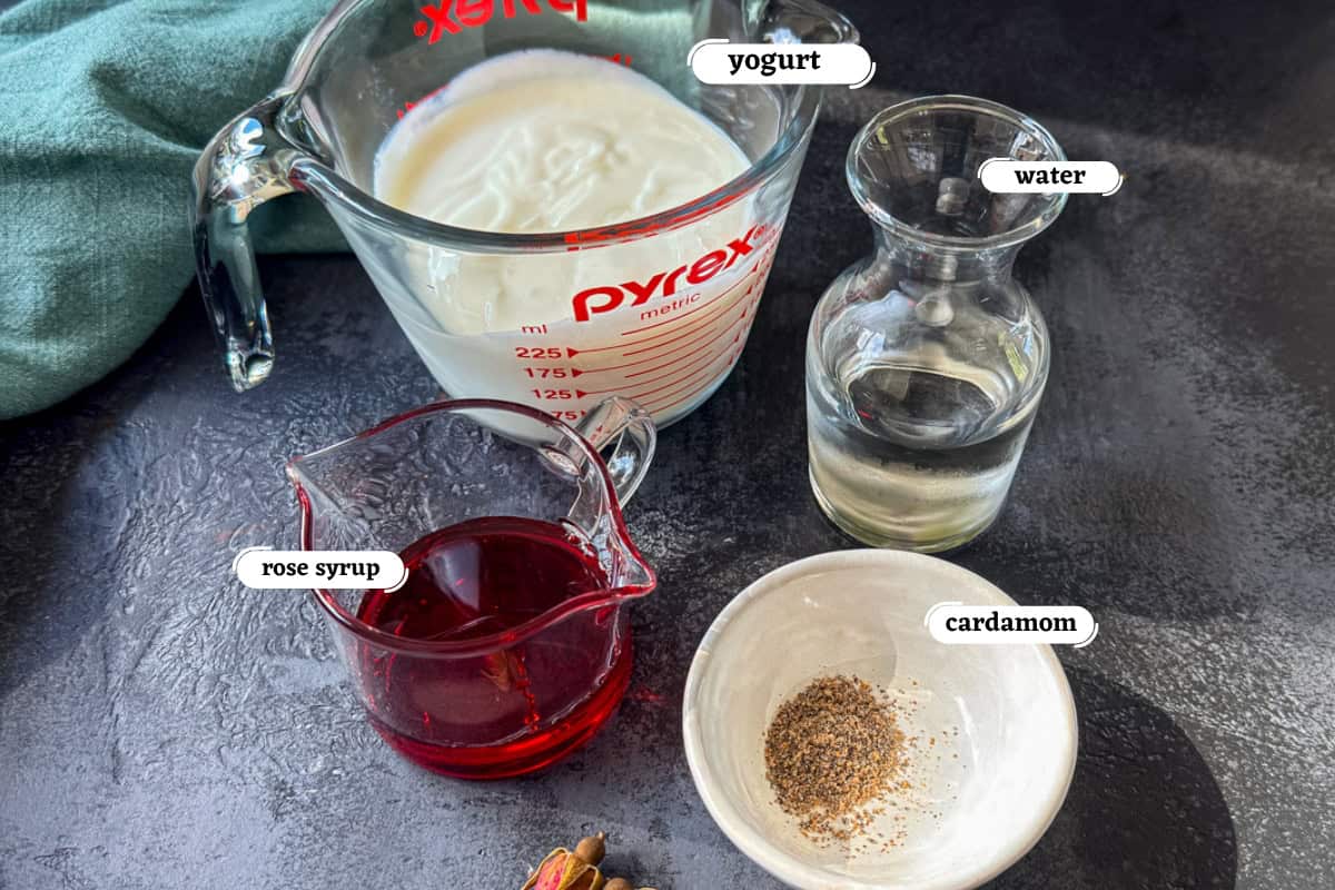 Yogurt, water, rose syrup, cardamom on grey board, ready to make a pink Indian yogurt drink called rose lassi