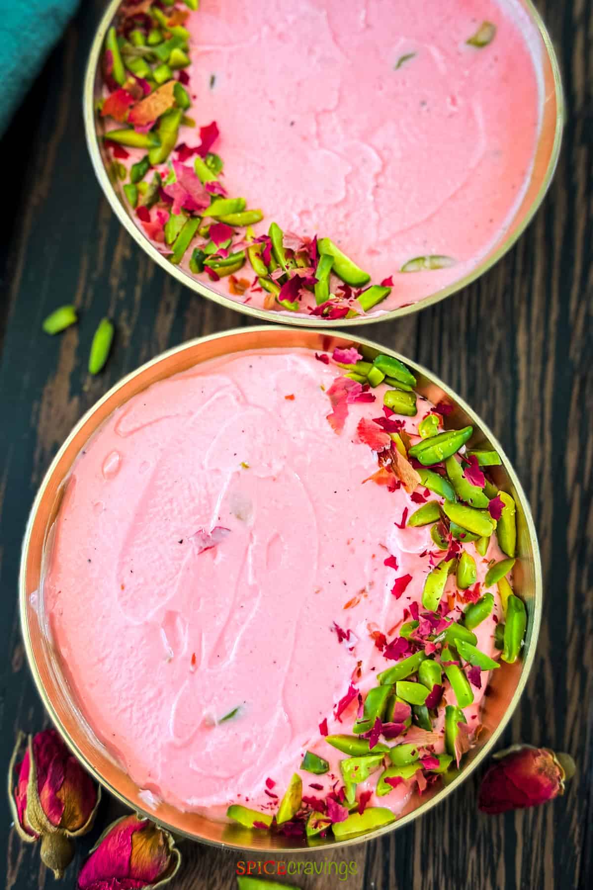 Overhead view of two brass bowls with creamy pink Rose Shrikhand garnished with slivered pistachios and dried rose petals on a dark wooden surface.