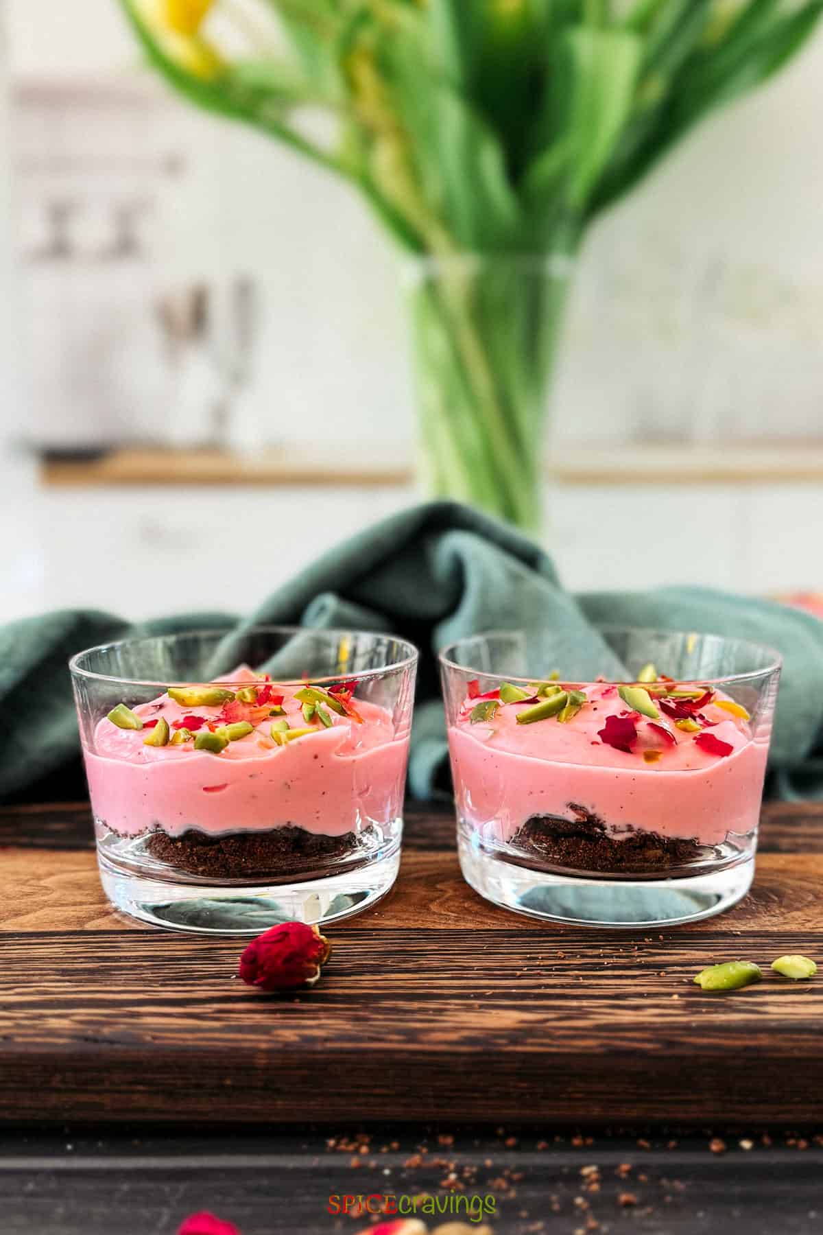 Two mini dessert bowls of creamy pink Rose Shrikhand garnished with slivered pistachios and dried rose petals on a dark chocolate cookie base.