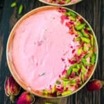 Overhead view of a brass bowl with pink Rose Shrikhand garnished with slivered pistachios and dried rose petals on a dark wooden surface.