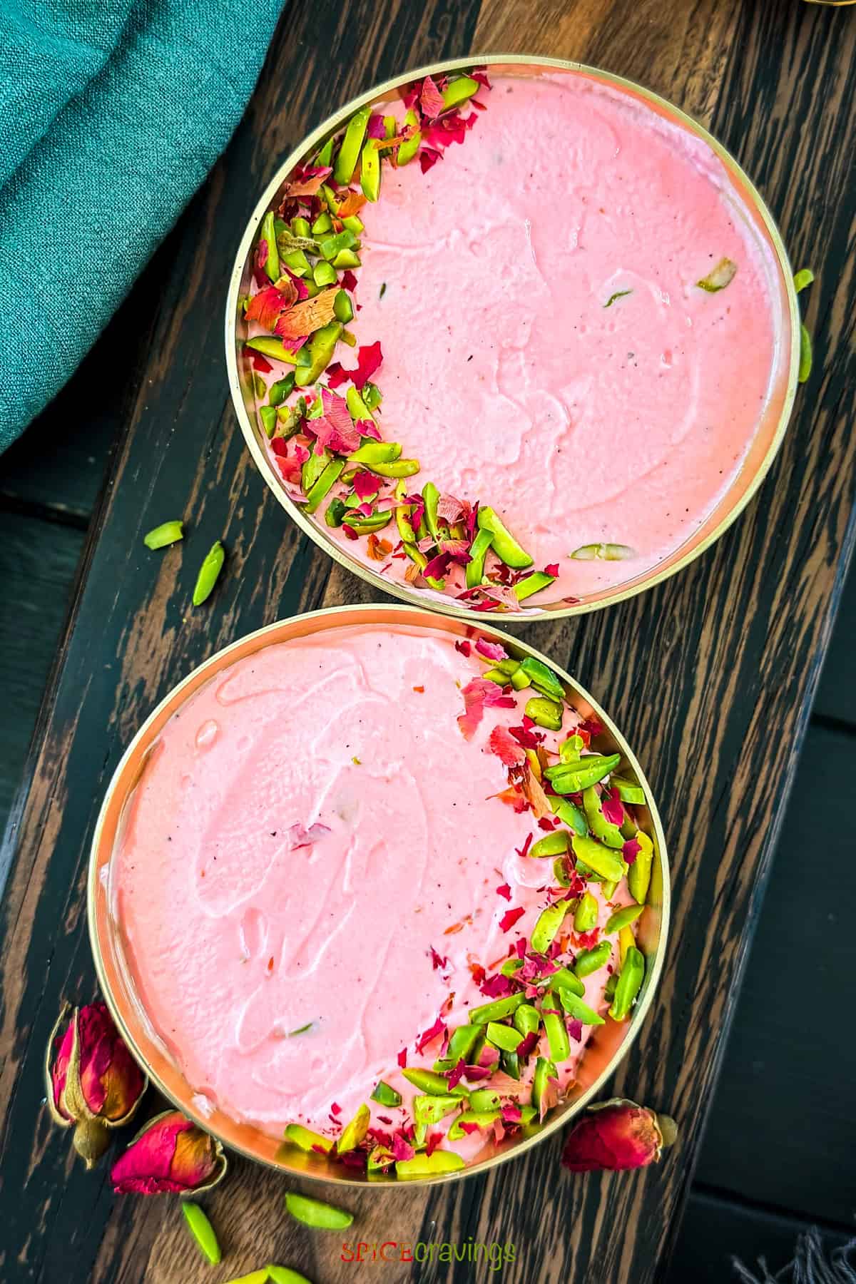 Two gold bowls of creamy pink Rose Shrikhand garnished with slivered pistachios and dried rose petals on a dark wooden surface.