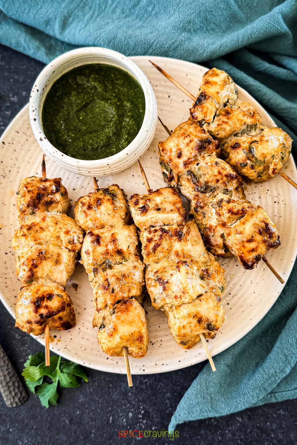 Five skewers of grilled Chicken Malai Tikka served on a cream speckled plate with a side of vibrant green cilantro chutney.