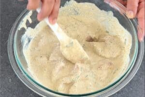 Cubed chicken breast being tossed in a creamy yogurt and spice marinade in a glass mixing bowl with a white spatula.