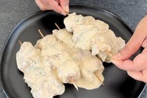 Hands threading marinated creamy chicken cubes onto wooden bamboo skewers over a black plate.