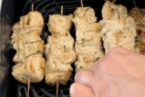 A hand flipping partially cooked chicken malai tikka skewers in an air fryer basket to ensure even cooking and charring.