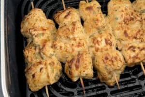 Fully cooked chicken malai tikka skewers in an air fryer basket with golden-brown charred edges and a glistening, creamy finish.